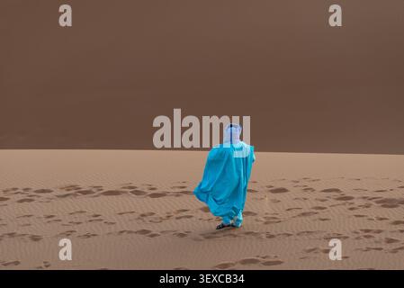 Bédouin en robes bleues marchant dans les dunes de sable du Sahara, Maroc. Ksebt An-Namous, Drâa-Tafilalet, Maroc. Les hommes et les femmes bédouins, en particulier ceux du Sahara et d'Afrique du Nord (souvent culturellement proches des Touaregs), portent des robes bleues amples et fluides distinctives appelées daraa ou boubou. Celles-ci sont associées à un long voile ou turban en tissu appelé tagelmust Banque D'Images