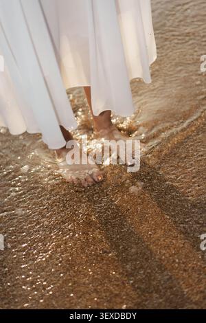 Femme pieds nus en robe blanche debout sur l'eau ondulations sur la plage de sable le matin Banque D'Images