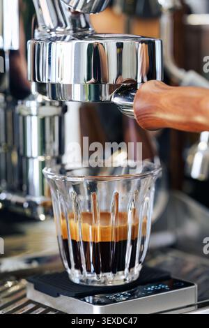 Une dose d'espresso riche s'écoulant dans une tasse en verre transparent positionnée sur l'échelle numérique sous le porte-filtre chromé poli avec poignée en bois sur l'esp professionnel Banque D'Images