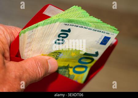 Main tenant enveloppe rouge remplie de billets de 100 euros, espèces cachées, salaire, cadeau, épargne Banque D'Images