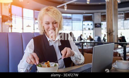 Femme d'affaires âgée caucasienne appréciant la salade tout en travaillant sur ordinateur portable dans le café confortable, sourire doux, vie professionnelle équilibrée Vibe, intérieur rempli de lumière Banque D'Images
