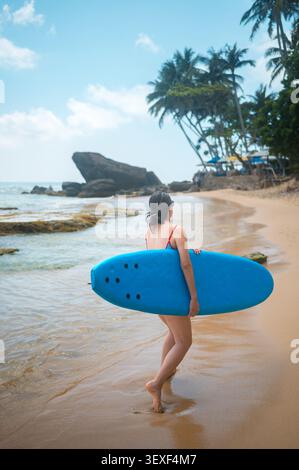 Une femme asiatique dans les années 30 se promène le long d'une plage sereine au Sri Lanka, portant une planche de surf bleue. La scène capture l'océan tranquille, les palmiers, et s. Banque D'Images