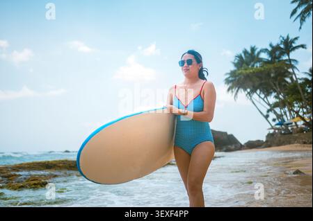 Une femme asiatique dans les années 30 se promène le long d'une plage pittoresque du Sri Lanka, tenant une planche de surf. Elle porte des lunettes de soleil et un maillot de bain élégant, avec un palmier Banque D'Images