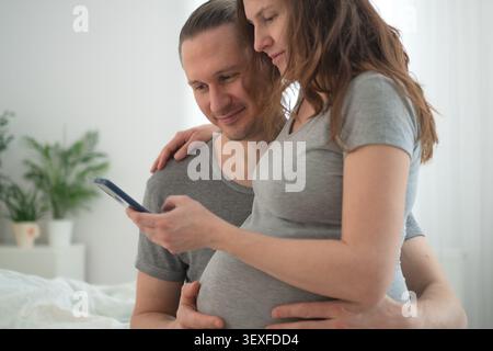 Femme enceinte et mari embrassant dans une chambre lumineuse partageant le jalon de la grossesse en privé. Attendant la mère avec le bras autour de la taille tandis que le partenaire tient le téléphone dans un moment intime calme ensemble. Banque D'Images