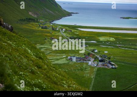Vue aérienne panoramique d'un petit village des Lofoten niché sur des pentes verdoyantes au-dessus des eaux turquoises de l'Arctique, avec des maisons traditionnelles, sinueuses Banque D'Images