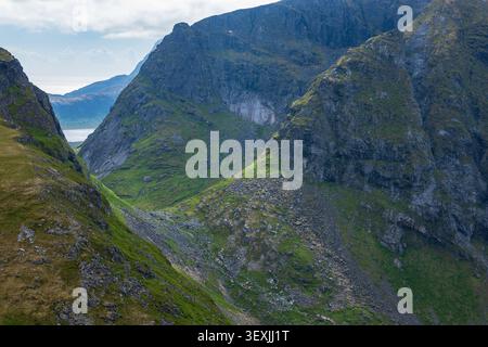 Vallée de montagne spectaculaire avec des falaises abruptes, des pentes vertes et un lac lointain de fjord dans les Lofoten, Norvège. Paysage alpin accidenté avec couches Banque D'Images