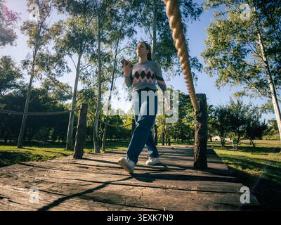 Femme marche sur un chemin en bois ensoleillé. Elle porte un pull coloré et un Jean bleu. De grands arbres verts entourent le sentier. La lumière du soleil filtre à travers le CA Banque D'Images