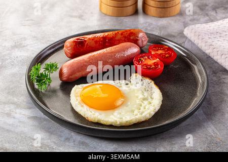Oeuf frit avec jaune liquide, deux saucisses grillées, tomates cerises coupées en deux disposées sur une plaque de céramique foncée. Garniture de persil frais. Éclairage naturel doux h Banque D'Images