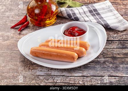Trois saucisses disposées sur une assiette blanche avec un petit bol de ketchup. Fond comprend bouteille d'huile de Chili, piments rouges, serviette à carreaux sur rustique Banque D'Images