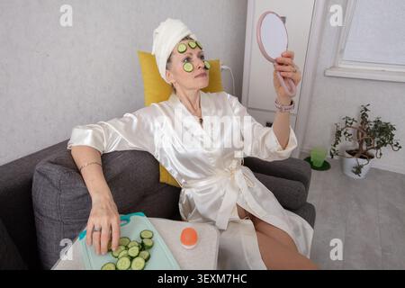 Une personne âgée vêtue de vêtements blancs, s'assoit confortablement sur un canapé gris, appliquant des tranches de concombre sur son visage quand elle regarde dans un petit miroir. Un tr Banque D'Images