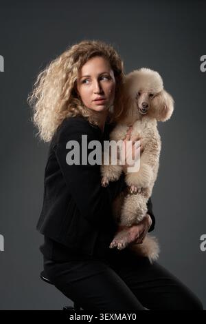 Une femme blonde embrasse un caniche blanc fermement tout en étant assise dans un studio sombre. Leur proximité et leurs expressions véhiculent un lien chaleureux et affectueux. Banque D'Images