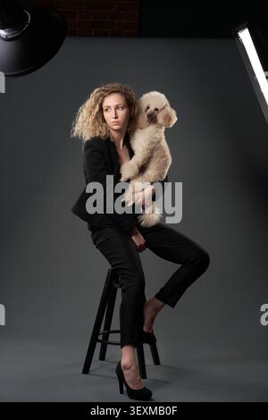 Un caniche blanc est tenu par une femme dans un photoshoot de mode avec éclairage de studio. La composition met l'accent sur l'élégance, la posture et la connexion. Banque D'Images