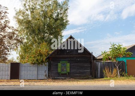Une petite vieille maison en bois avec une fenêtre verte se dresse devant une clôture. La maison est entourée d'arbres et de buissons. Banque D'Images