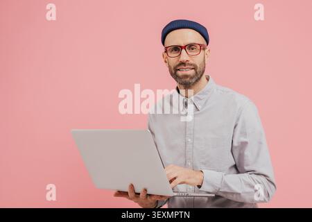 Photo de Guy internaute navigue sur le site Web sur ordinateur portable, connecté à Internet sans fil, tient un gadget électronique, porte des lunettes, chemise formelle, isolé sur fond rose avec de l'espace libre Banque D'Images