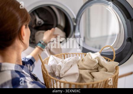 La femme charge des vêtements légers du panier à linge en osier dans la machine à laver à chargement frontal. Banque D'Images