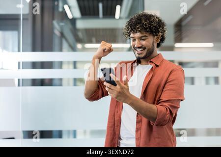 Jeune homme debout dans un bureau moderne, regardant joyeusement son smartphone et fermant un poing, exprimant de la joie et célébrant une réussite en ligne, une victoire ou une bonne nouvelle Banque D'Images