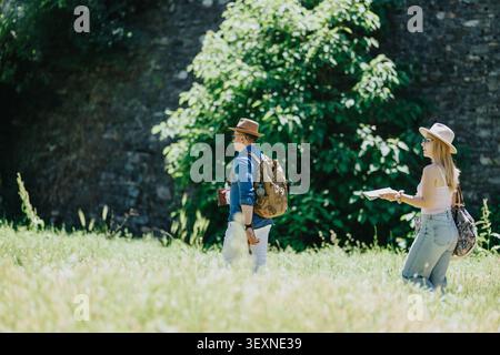 Couple de touristes marchant à travers un champ ensoleillé avec des sacs à dos et une carte près d'un mur de pierre Banque D'Images