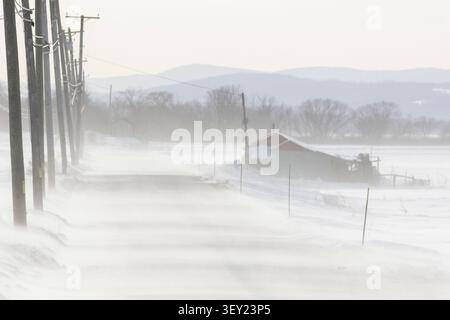 Les vents de Strongs soufflent la neige des champs agricoles à travers une route en hiver. Banque D'Images