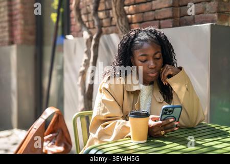 Femme afro-américaine réfléchie en pause café dans un café, assise à la table défilant sur smartphone Banque D'Images