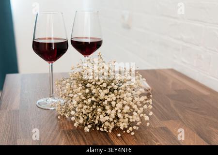 Une composition romantique avec deux élégants verres remplis de vin rouge positionnés à côté d'un délicat bouquet de fleurs blanches séchées de l'haleine de bébé, Banque D'Images