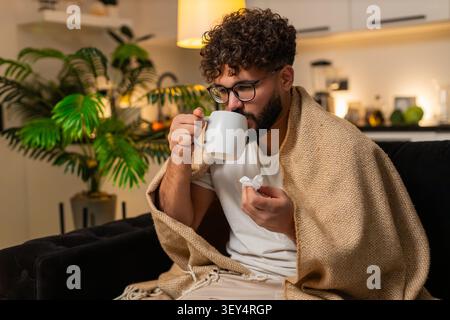 L'homme arabe à la maison se sent mal à la gorge tient tasse de thé chaud avec la médecine prend lente gorgée utilise le tissu. Égyptien sur le canapé fouille médecin sur smartphone frotte toux cou repose sous la fièvre sniff couverture Banque D'Images