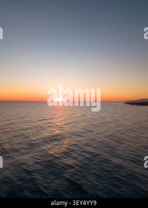 La perspective de drone à angle élevé capture un soleil brillant se levant sur la surface calme de l'océan près d'une ville côtière lointaine à l'aube. Banque D'Images