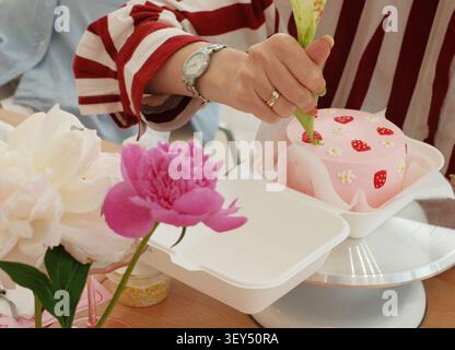 Femme en pull rayé à l'aide d'un sac à passepoil pour décorer un mini gâteau bento rose avec des fraises fondantes et des marguerites sur un support à gâteau, pivoines roses Banque D'Images