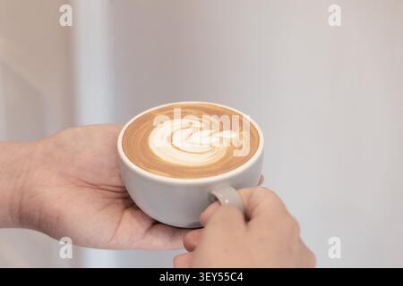 Close-up de deux mains tenant une tasse en céramique grise de latte remplie d'art au mousse de lait en spirale, posée sur un fond clair et propre. Banque D'Images