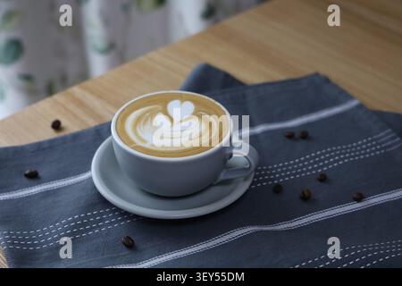 Une tasse de latte ornée d'un cœur et d'une tulipe en mousse de lait repose sur un tissu à carreaux sombres avec des grains de café éparpillés sur une table en bois, photographiée depuis un angle latéral légèrement surélevé. Banque D'Images
