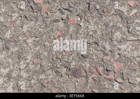 Une vue rapprochée d'une surface grossière en béton altérée avec des pierres rose rougeâtre de différentes tailles et des cailloux encastrés dans le ciment gris. Banque D'Images