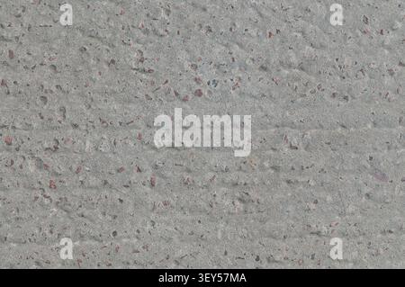 Une vue rapprochée d'une surface de béton rugueuse et grise avec de petits fragments de pierre rouge et brun encastrés. La texture est inégale et d'aspect industriel. Banque D'Images