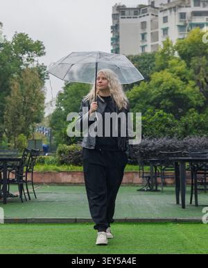 Jeune femme aux cheveux blonds se tient sous un parapluie clair pendant une douche de pluie de jour Banque D'Images