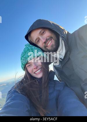 Couple souriant en équipement de plein air prend selfie sur le sommet de la montagne pendant la randonnée. Visages heureux de jeune homme et femme en vêtements chauds, joyeux moment de shar Banque D'Images