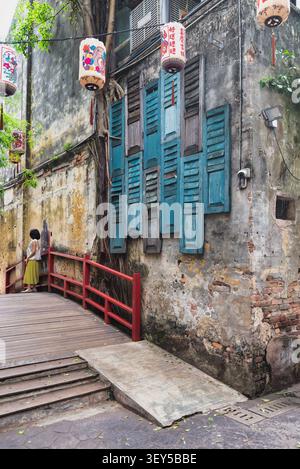 Femme sur un pont en bois avec des garde-corps rouges dans l'allée de Kwai Chai Hong, Kuala Lumpur, Malaisie. Ruelle historique avec murs altérés, volets bleus et lanternes chinoises vibrantes Banque D'Images