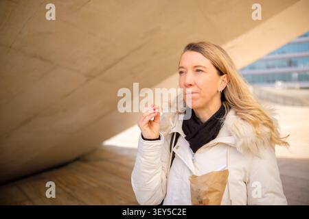 Femme blonde profitant d'une collation dans un environnement urbain à l'architecture contemporaine Banque D'Images