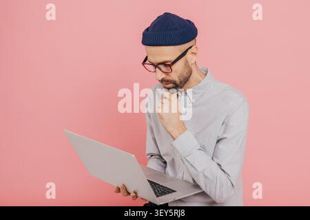 Indépendant masculin expérimenté développe une nouvelle stratégie, tient un ordinateur portable moderne, garde la main sur le menton, porte des vêtements formels, occupé avec le travail à distance, isolé sur fond rose, espace de copie. Banque D'Images