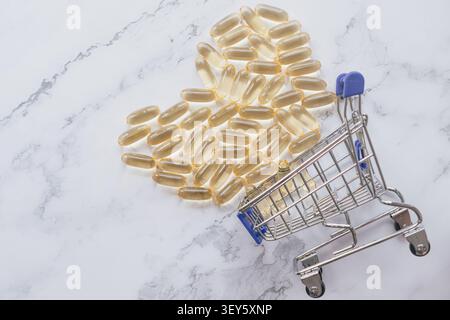 Plan horizontal à grand angle de capsules d'huile de poisson oméga dorées disposées en forme de coeur avec un petit chariot sur un fond de marbre blanc sous b. Banque D'Images