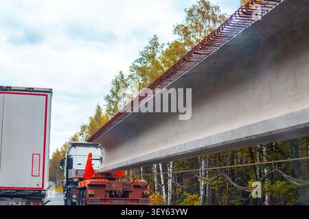 Un camion lourd transporte une poutre de pont massive en béton sur une autoroute en construction. transport de marchandises industrielles, ingénierie lourde, pont bui Banque D'Images