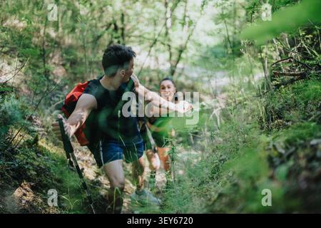 Jeune couple randonnant dans un sentier forestier vert avec des sacs à dos et des bâtons de randonnée s'aidant les uns les autres Banque D'Images