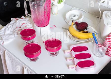 Mélanger les fraises, les bananes, les betteraves et le yogourt grec dans un mélangeur pour créer de la crème glacée. Les bols et les moules à glaçons reposent sur un comptoir de cuisine avec Banque D'Images