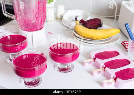 Mélanger les fraises, les bananes, les betteraves et le yogourt grec dans un mélangeur pour créer de la crème glacée. Les bols et les moules à glaçons reposent sur un comptoir de cuisine avec Banque D'Images