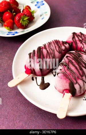 Trois popsicles à base de fraises, de bananes, de betteraves et de yaourt grec sont posées sur une assiette. Ils sont arrosés de chocolat et dégustés pendant un d ensoleillé Banque D'Images
