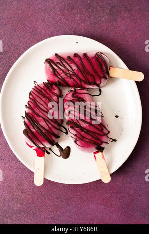 Trois popsicles à base de fraises, de bananes, de betteraves et de yaourt grec sont posées sur une assiette. Ils sont arrosés de chocolat et dégustés pendant un d ensoleillé Banque D'Images