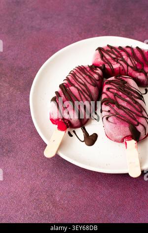 Trois popsicles à base de fraises, de bananes, de betteraves et de yaourt grec sont posées sur une assiette. Ils sont arrosés de chocolat et dégustés pendant un d ensoleillé Banque D'Images