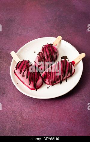 Trois popsicles à base de fraises, de bananes, de betteraves et de yaourt grec sont posées sur une assiette. Ils sont arrosés de chocolat et dégustés pendant un d ensoleillé Banque D'Images
