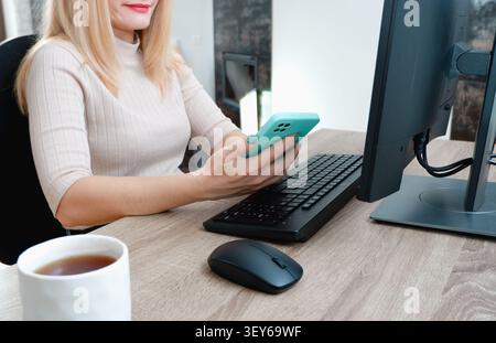 Femme d'affaires tenant un smartphone dans ses mains tout en travaillant de la maison à l'ordinateur. Télétravail. Mise au point sélective. Mains, souris, clavier, blanc Banque D'Images