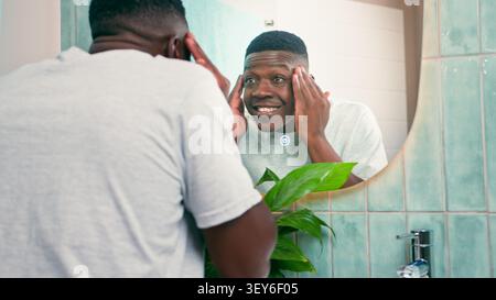 Jeune homme noir souriant à Mirror tout en pratiquant les soins de la peau dans la salle de bain lumineuse avec Plant Banque D'Images