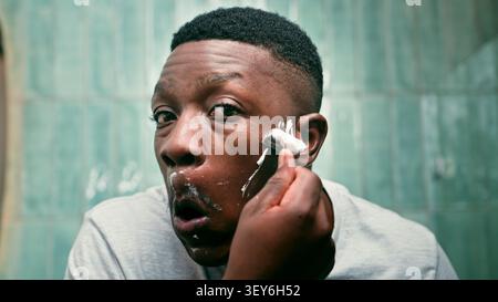 Jeune homme rasant avec rasoir dans la salle de bains miroir routine de toilettage matin gros plan Banque D'Images