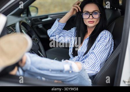 Un modèle tendance brune avec des lunettes se trouve confortablement dans une cabine de voiture moderne avec ses pieds en haut. Elle porte une chemise rayée bleue et un Jean vieilli, Banque D'Images