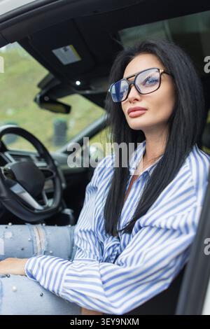 Vue latérale d'une jeune conductrice à la mode reposant à l'intérieur d'une cabine de voiture moderne. Portant une blouse rayée bleue et un denim vieilli avec des perles, elle Banque D'Images